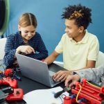 Three children collaborating around a laptop with a 3D printer and robotic builds at a robotics camp