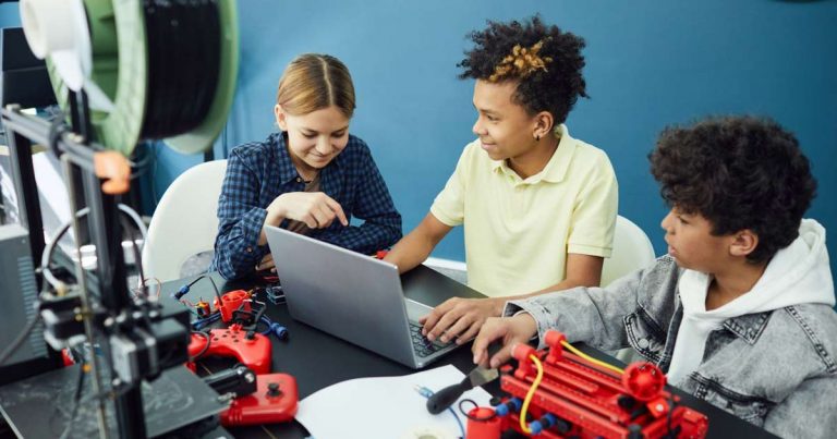 Three children collaborating around a laptop with a 3D printer and robotic builds at a robotics camp