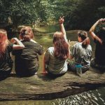 Group of young people sitting on a log by a forest pool surrounded by lush green trees