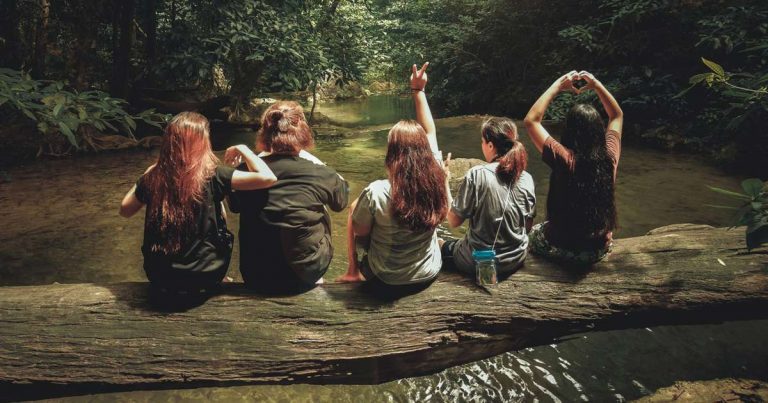 Group of young people sitting on a log by a forest pool surrounded by lush green trees