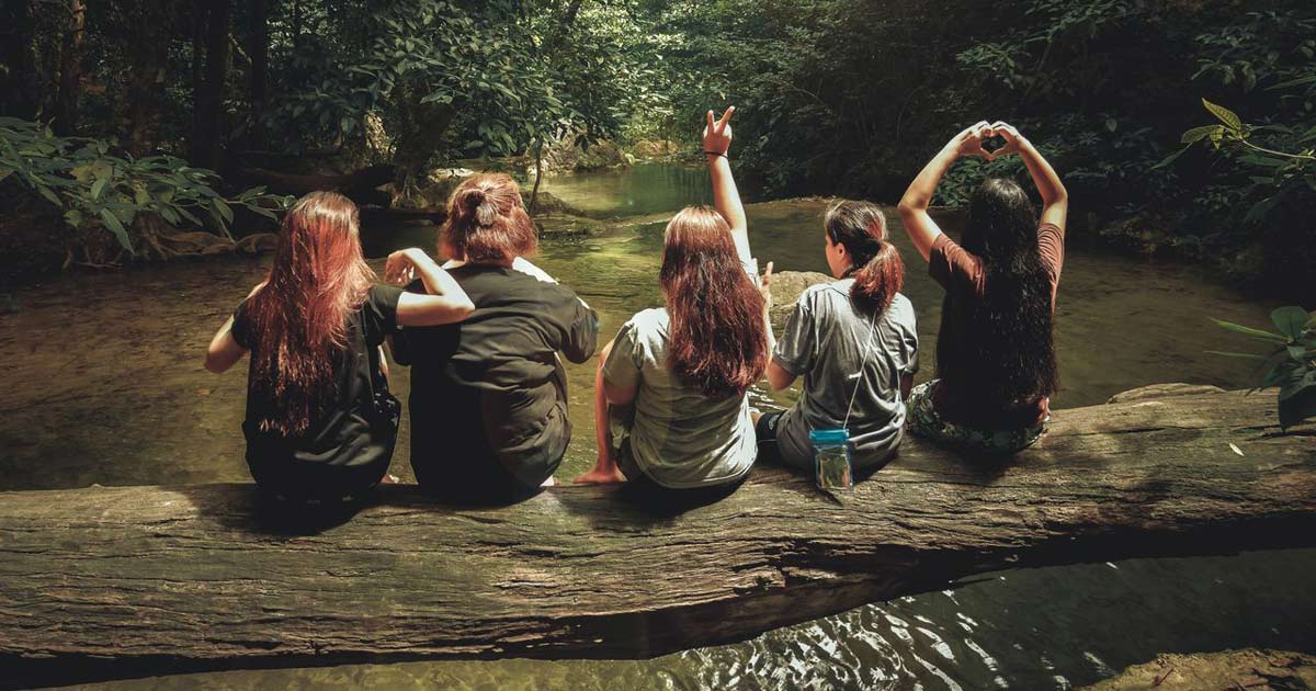Group of young people sitting on a log by a forest pool surrounded by lush green trees