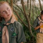 Two children in camp uniforms outdoors in a woodland setting, one alert and engaged, one writing in a notebook