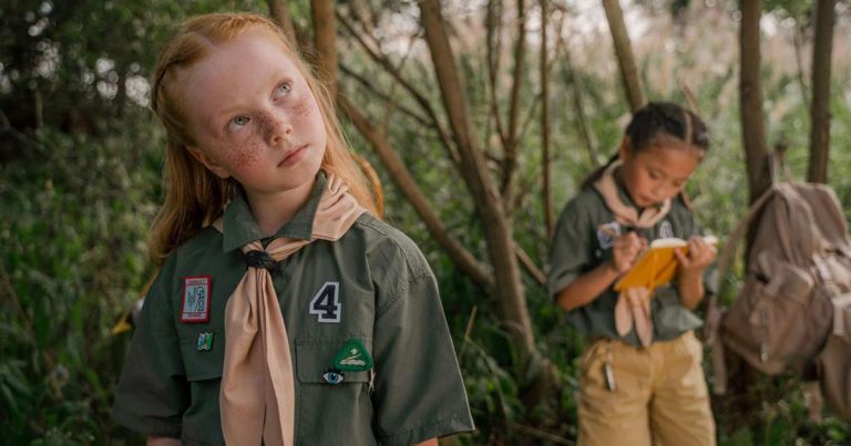 Two children in camp uniforms outdoors in a woodland setting, one alert and engaged, one writing in a notebook
