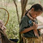 Child in camp uniform writing in a notebook outdoors among trees and camp gear