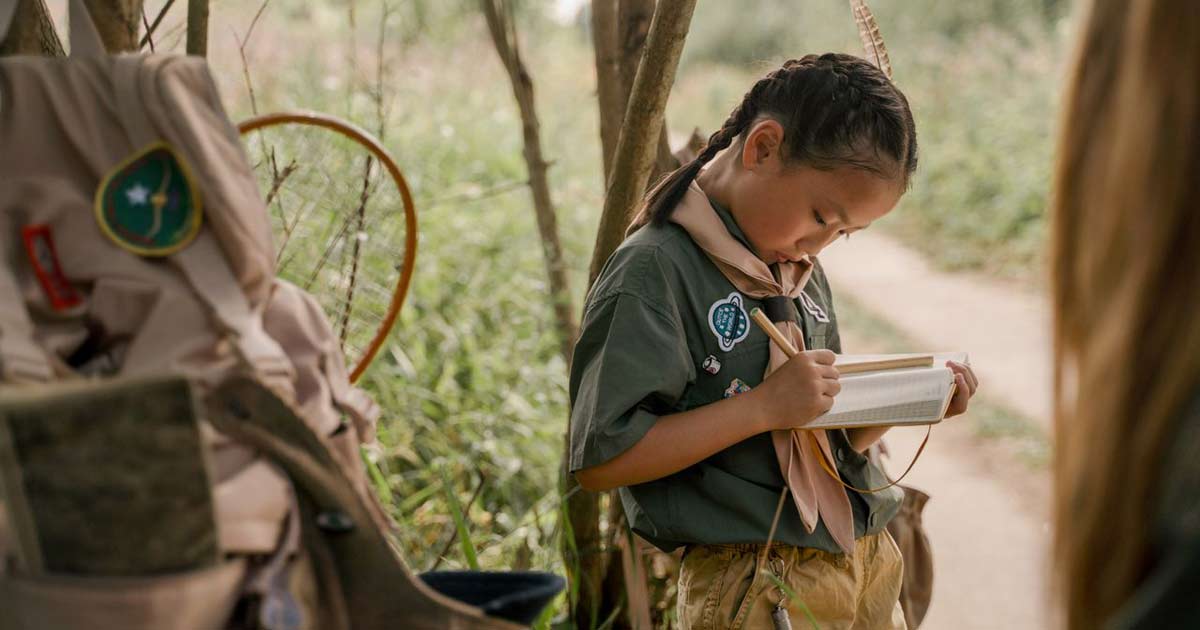 Child in camp uniform writing in a notebook outdoors among trees and camp gear