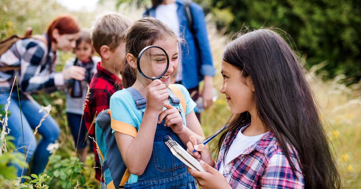 Children using a magnifying glass and notebook during a guided nature study activity with counselors at summer camp