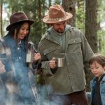 Family of four standing together among the trees at a family camp holding mugs in the morning mist