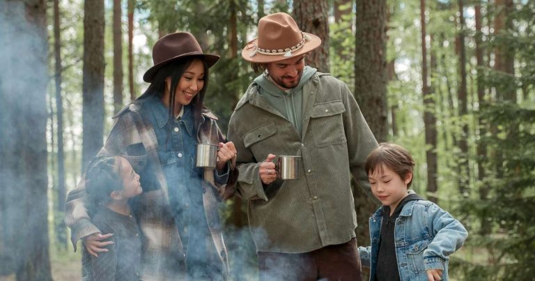 Family of four standing together among the trees at a family camp holding mugs in the morning mist