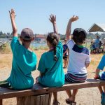 Group of children seated on a bench raising their hands during a waterfront summer camp activity