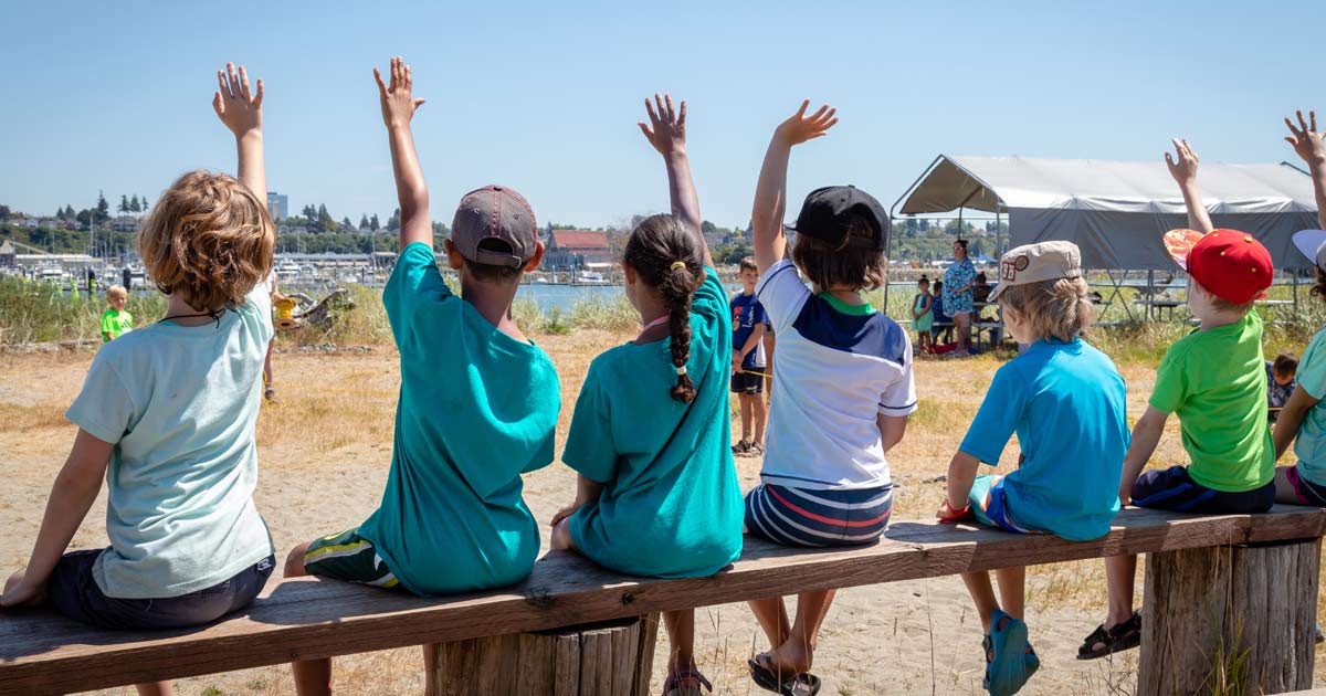 Group of children seated on a bench raising their hands during a waterfront summer camp activity