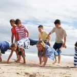 Group of children playing a wheelbarrow race game on the beach at summer camp