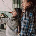Mother guiding her daughter's aim during an archery activity at a parent-child camp