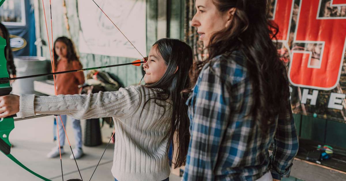 Mother guiding her daughter's aim during an archery activity at a parent-child camp