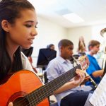 Teenage girl playing acoustic guitar from sheet music in a group music class