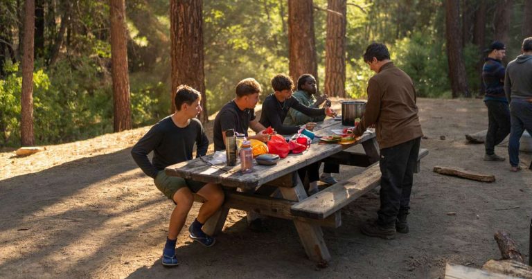 Camp staff member serving food to a group at an outdoor picnic table in the woods