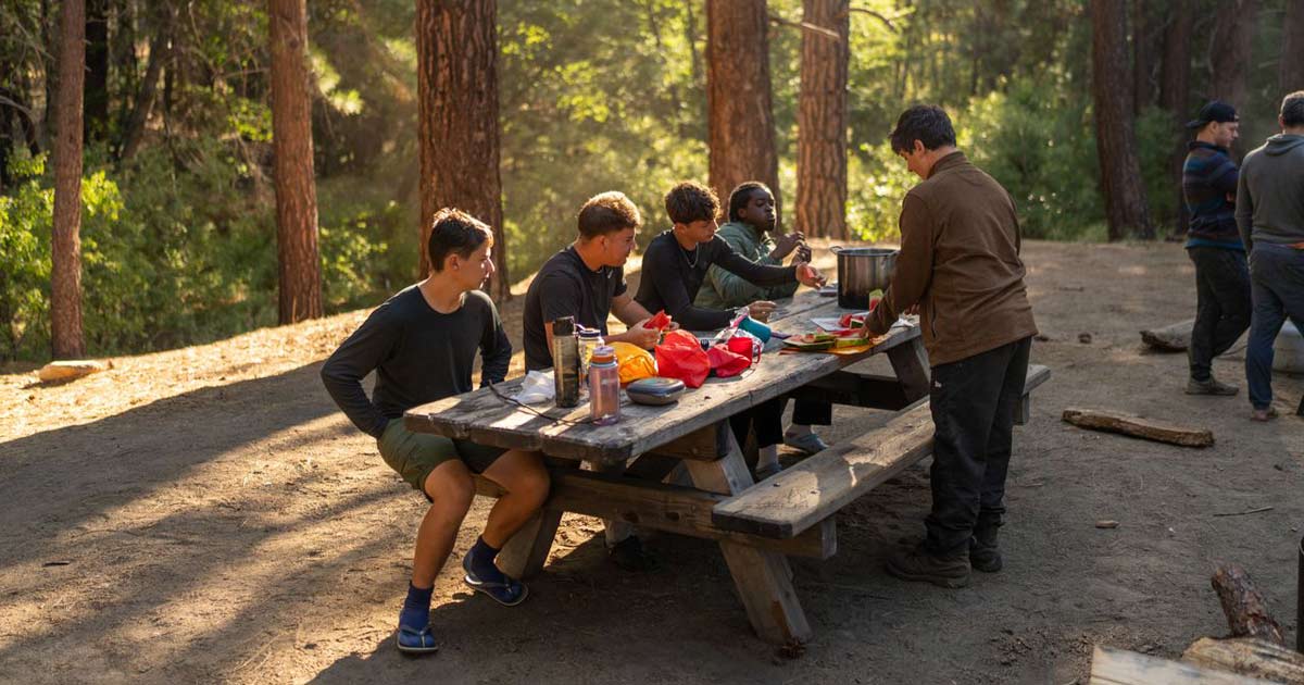 Camp staff member serving food to a group at an outdoor picnic table in the woods