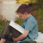 Boy sitting on a blanket by a lake at summer camp reading quietly in the evening light