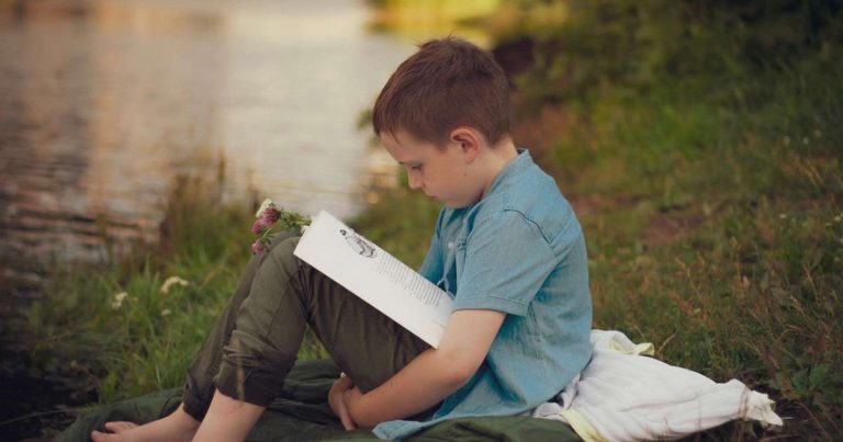 Boy sitting on a blanket by a lake at summer camp reading quietly in the evening light