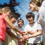 Five children stacking hands together outdoors under a blue summer sky