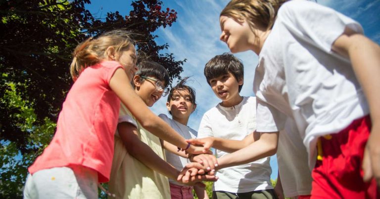 Five children stacking hands together outdoors under a blue summer sky