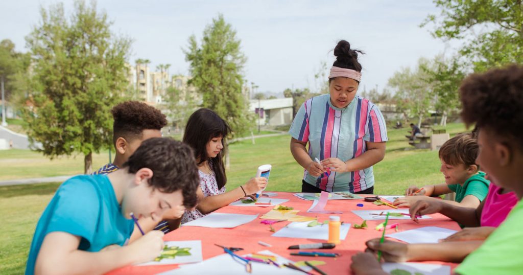 Young adult camp counselor leading an outdoor arts activity with a group of campers