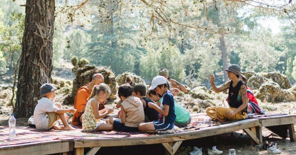 Group of children and an adult seated together on a wooden outdoor platform under a large pine tree
