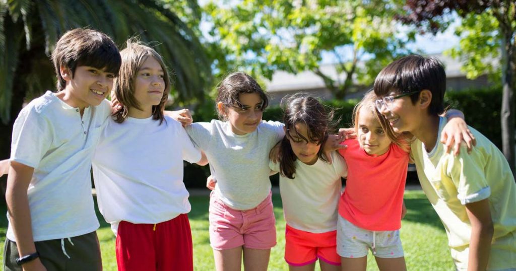 Six campers with arms around each other's shoulders, smiling outdoors at camp